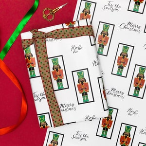 May include: White wrapping paper with a repeating pattern of a nutcracker soldier in a black frame, with the phrases "Merry Christmas", "Happy Holidays", and "Tis the Season". A gift is wrapped with a brown ribbon with red and green polka dots.