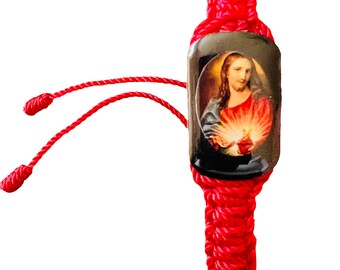 May include: Red string bracelet with a small rectangular charm featuring a religious image of Jesus Christ with a glowing heart.