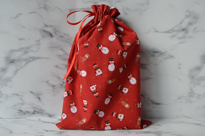 May include: Red fabric gift bag with a drawstring closure and a red ribbon. The bag is decorated with a repeating pattern of snowmen and reindeer.