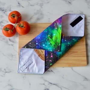 May include: A reusable food wrap with a galaxy print design. The wrap is folded over a slice of bread. The wrap is made of fabric and has a velcro closure.