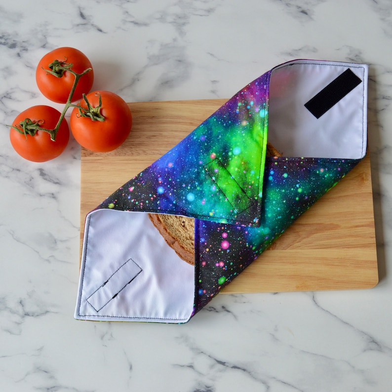 May include: A reusable food wrap with a galaxy print design. The wrap is folded over a slice of bread. The wrap is made of fabric and has a velcro closure.