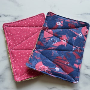 May include: Two quilted pot holders, one with a pink and white polka dot pattern and the other with a blue and pink floral pattern. Both pot holders have a hessian border.