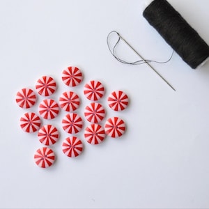May include: A selection of red and white striped buttons, reminiscent of peppermint sweets, arranged on a white background. A black reel of thread and a needle with black thread are also visible, indicating sewing or craft work.