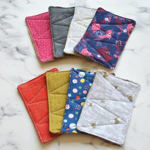 May include: Eight rectangular fabric coasters in assorted colours and patterns. Designs include polka dots, floral, geometric, and bee motifs. The coasters are quilted with a stitched border.