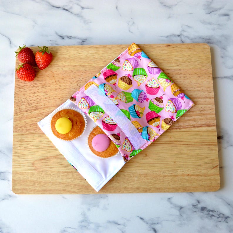 Pink reusable snack bag with a cupcake design on a wooden bread board.  The snack bag has white hook and loop tape the secure the food inside.