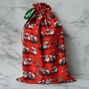 May include: Red drawstring gift bag with a Christmas pattern featuring snowmen, reindeer, and trees. The bag has a green ribbon.