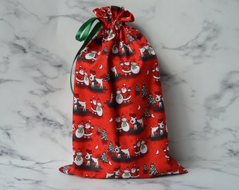 May include: Red drawstring gift bag with a Christmas pattern featuring snowmen, reindeer, and trees. The bag has a green ribbon.