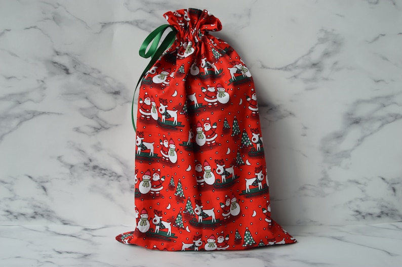 May include: Red drawstring gift bag with a Christmas pattern featuring snowmen, reindeer, and trees. The bag has a green ribbon.