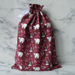 May include: A burgundy drawstring gift bag with a white polar bear and penguin Christmas print. The bag has a white drawstring.