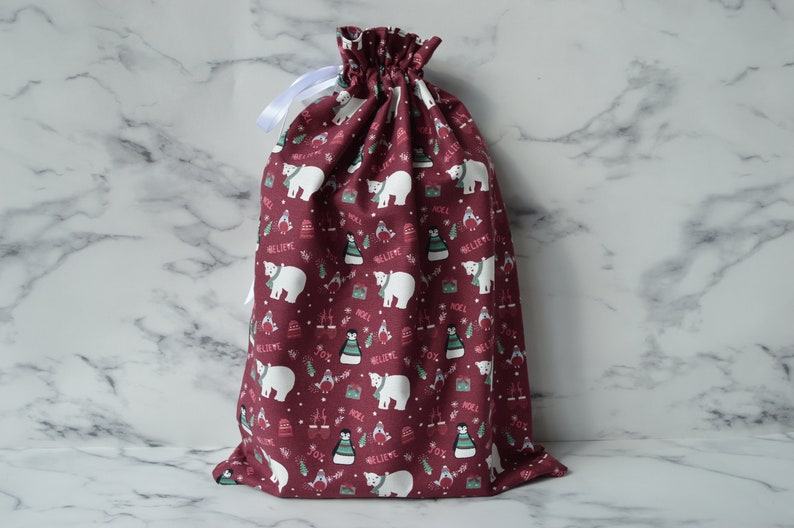 May include: A burgundy drawstring gift bag with a white polar bear and penguin Christmas print. The bag has a white drawstring.