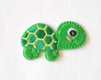 Puede incluir: Un parche bordado de una tortuga verde con un patrón hexagonal amarillo en el caparazón. La tortuga tiene una cabeza redondeada con un ojo visible, una pequeña boca rosada y un borde cosido alrededor de toda la figura.