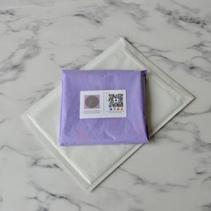May include: A purple padded envelope with a white label containing a small circle with a needle and thread inside and a QR code with social media icons below it.
