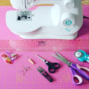May include: A pink cutting mat with a white sewing machine, a ruler, a pair of scissors, a rotary cutter, and other sewing supplies.