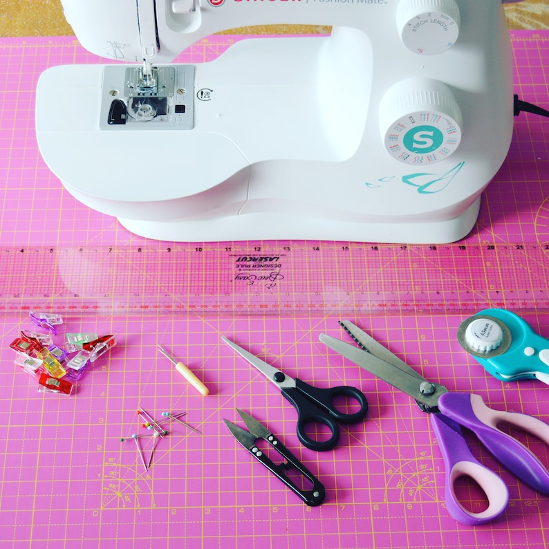 May include: A pink cutting mat with a white sewing machine, a ruler, a pair of scissors, a rotary cutter, and other sewing supplies.