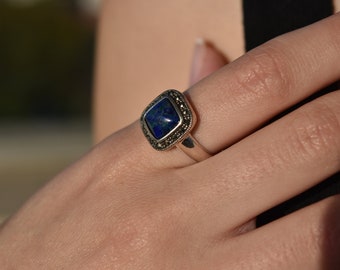 May include: A silver ring featuring a square, blue gemstone with green and gold flecks. The gemstone is set in a square frame with small, dark accents. The ring is on a finger.