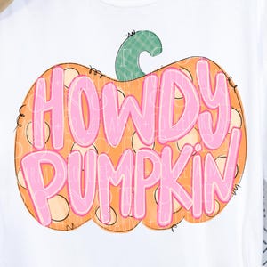 May include: White t-shirt with a pumpkin graphic that says "HOWDY PUMPKIN" in pink letters. The pumpkin is orange with a green stem. A round logo with flowers and text is in the lower right corner.