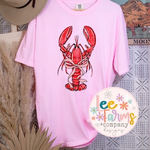 May include: Pink t-shirt with a red lobster illustration wearing a pink bow. The lobster is centered on the shirt. The shirt has short sleeves and a crew neck.