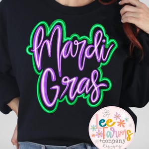 Op de afbeelding: Een zwart sweatshirt met een neon groene en paarse grafische afbeelding met de tekst "Mardi Gras". Het sweatshirt wordt gedragen door een persoon met lang rood haar. Een kleine roze cirkel met de tekst "Lee Farms + Company Designs" en een bloemenontwerp bevindt zich in de rechterbenedenhoek van de afbeelding.