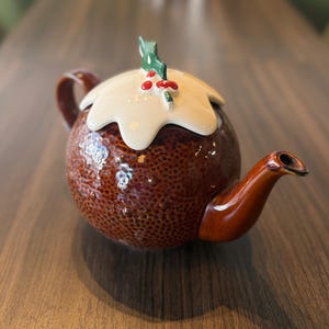 Op de afbeelding: Een bruine keramische theepot met een ronde vorm, een getextureerd oppervlak, een crèmekleurig deksel en een bruine handgreep en tuit. Het deksel is versierd met groene hulstbladeren en rode bessen. De theepot staat op een houten tafel.