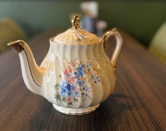 Può includere: Teiera in ceramica color crema con un design floreale rosa e blu. La teiera ha un manico, un beccuccio e dettagli dorati sul coperchio. Il corpo della teiera presenta delle scanalature verticali. Un pezzo classico per servire il tè.