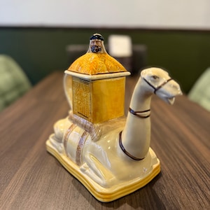 May include: A ceramic teapot shaped like a camel with a yellow and brown patterned design. The camel is carrying a small, yellow, square-shaped container with a man's face on the lid.