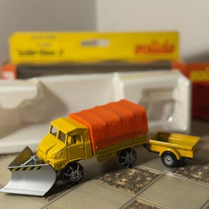 May include: A yellow toy truck with an orange cargo cover and a silver snowplow attached. The truck is pulling a yellow trailer. The wheels have black tire chains. The background includes a yellow box.