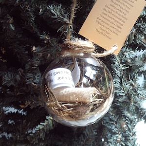 May include: A clear glass ornament with a brown twine hanger. Inside the ornament is straw, a small piece of white fabric, and a white paper tag with the text "He gave His life. John 3:16". A brown paper tag with a poem about the birth of Jesus is attached to the ornament.