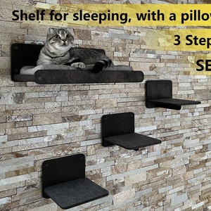 May include: A three-tiered cat shelf set made of black wood with a gray cushion on the top shelf. A cat is lying on the top shelf. The text "Shelf for sleeping, with a pillow. 3 Steps. SET." is visible on the wall.