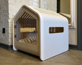May include: White wooden dog house with a clear plastic door and a brown cushion inside. The house has a rounded roof and a small window on the side.