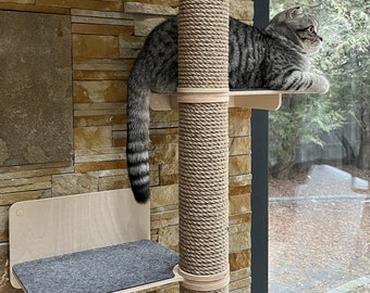 May include: A gray tabby cat sits on a wooden platform attached to a sisal-wrapped scratching post. The cat is looking out a window. The scratching post is mounted to a wall with a stone veneer.