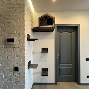 Puede incluir: Una pared de ladrillo blanco con un árbol para gatos fijado a ella. El árbol para gatos tiene una casa, una plataforma y algunas estanterías. El árbol para gatos está hecho de madera y tiene un acabado marrón oscuro. Hay una puerta a la derecha del árbol para gatos.