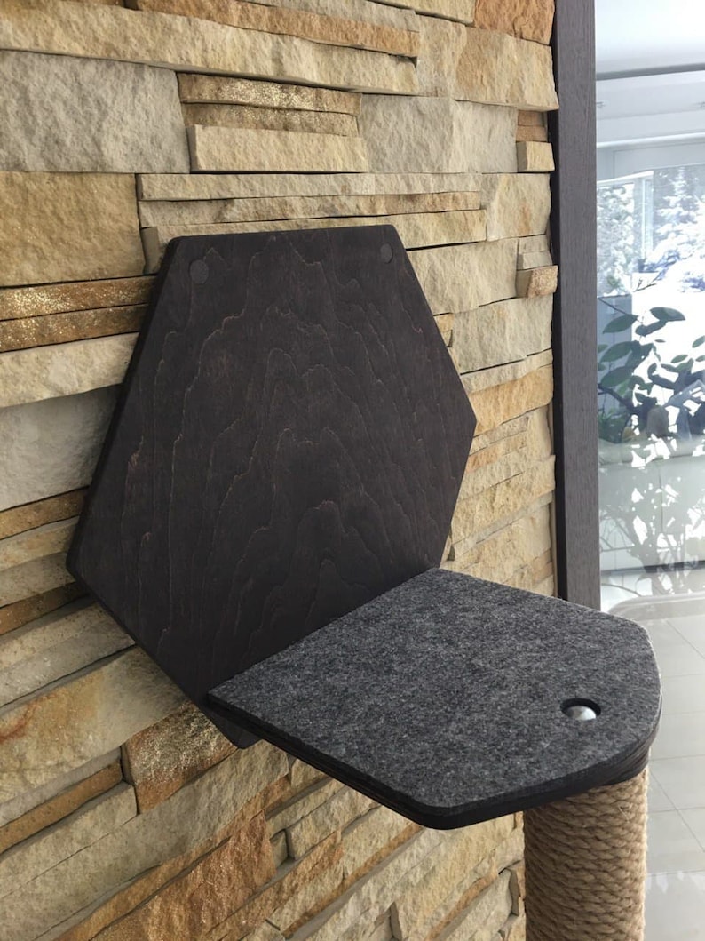 May include: A dark brown wooden cat perch with a gray felt platform and a sisal rope post. The perch is mounted to a stone wall.