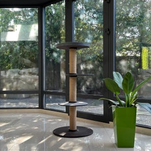 May include: A dark gray cat tree stands on a light-colored tile floor in front of a large window. The cat tree has three circular platforms connected by a thick, tan sisal rope post. The top and bottom platforms are larger than the middle platform. A green potted plant sits to the right of the cat tree. The window shows lush green trees and landscaping outside.