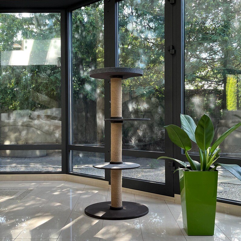 May include: A dark gray cat tree stands on a light-colored tile floor in front of a large window. The cat tree has three circular platforms connected by a thick, tan sisal rope post. The top and bottom platforms are larger than the middle platform. A green potted plant sits to the right of the cat tree. The window shows lush green trees and landscaping outside.