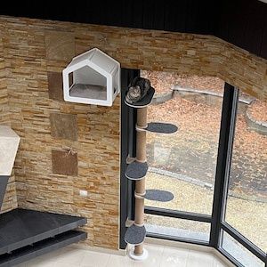 K&ouml;nnte beinhalten: Ein wei&szlig;er Katzenbaum mit einer grauen Plattform und einem wei&szlig;en Katzenbett, das an der Wand h&auml;ngt. Der Katzenbaum ist aus Sisal-Seil gefertigt und hat eine wei&szlig;e Basis. Das Katzenbett ist hausf&ouml;rmig und hat eine transparente Plastikfront.