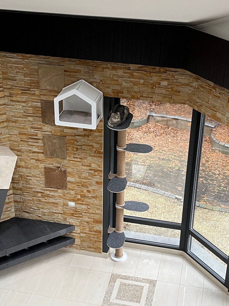 K&ouml;nnte beinhalten: Ein wei&szlig;er Katzenbaum mit einer grauen Plattform und einem wei&szlig;en Katzenbett, das an der Wand h&auml;ngt. Der Katzenbaum ist aus Sisal-Seil gefertigt und hat eine wei&szlig;e Basis. Das Katzenbett ist hausf&ouml;rmig und hat eine transparente Plastikfront.
