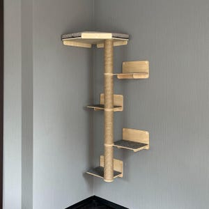 Op de afbeelding: Een houten kattenboom ontworpen voor een hoek. Het heeft een centrale sisal-omwikkelde paal met meerdere plateaus en een bovenste ligplaats met een grijs kussen. De plateaus hebben grijze viltpads.