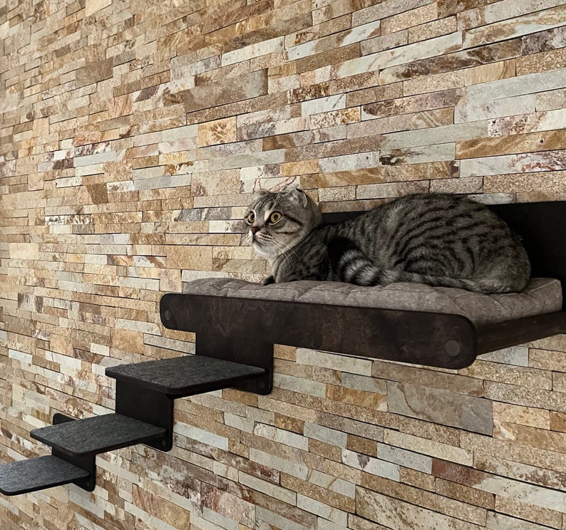May include: A gray tabby cat with green eyes sits on a black cat tree with a padded platform and steps. The cat tree is mounted on a wall with a stone-like texture.