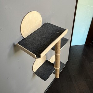 Puede incluir: Un &aacute;rbol para gatos de pared con tres plataformas cubiertas de tela gris y un poste para rascar de sisal. Las plataformas son de madera y tienen bordes redondeados.