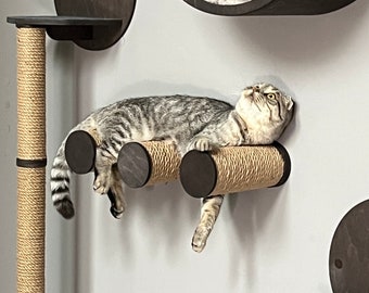 May include: A gray and white cat is lying on a cat scratching post made of sisal rope and wood. The scratching post is mounted on a white wall.