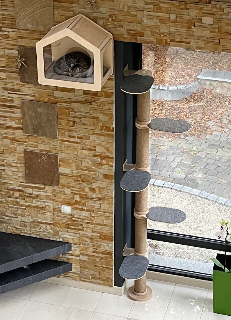 K&ouml;nnte beinhalten: Ein Katzenbaum mit einem beigefarbenen, mit Seil umwickelten Pfosten und vier grauen Plattformen. Der Katzenbaum ist an einer Wand neben einem Fenster montiert. Eine Katze schl&auml;ft in einem beigefarbenen Katzenhaus, das an der Wand &uuml;ber dem Katzenbaum montiert ist.
