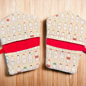 May include: Two red and white oven mitts with a pattern of cartoon milk cartons with faces. The mitts have a red lining and a red strap.