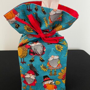May include: A blue and red fabric tissue box cover with a gnome and autumn theme. The cover has a red ribbon tied around it and features gnomes, birds, sunflowers, and pumpkins.