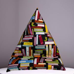 May include: A black triangular pillow with a colorful book print. The pillow is covered in a variety of colorful books with titles like "Dictionary", "History", and "Science".