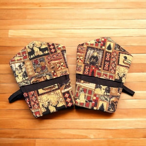 May include: A pair of brown and beige oven mitts with a woodland pattern featuring deer, bears, and plaid designs. The mitts have black straps and are made of fabric.
