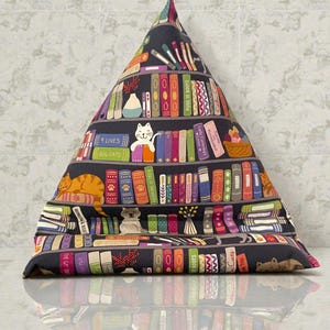 May include: A dark gray, triangular pillow with a bookshelf design. The pillow features colorful books, cats, and the text "9 Lives" and "Big Cats." The design includes the text "Puss in Boots" and "Lady Catterley's Lover."