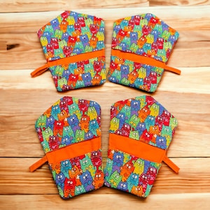 May include: Four colorful oven mitts with a pattern of cartoon cats. The mitts are orange, green, blue, and purple with a bright orange trim.