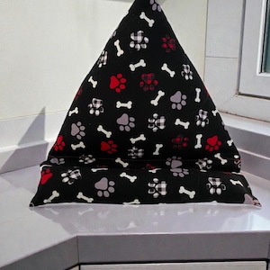 May include: A black fabric tablet stand with a triangular shape, patterned with white bones, red and grey paw prints. The stand is designed to prop up a tablet or e-reader.