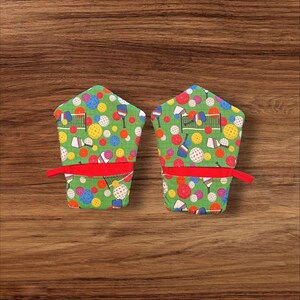 May include: A pair of green oven mitts with a red trim. The mitts are decorated with a pattern of colorful pickleball paddles, balls, and nets.
