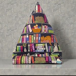 Peut inclure: Un support de livre en forme de pyramide avec un motif d'étagère. Le tissu présente des livres colorés, des chats et un panier de laine. La base est rectangulaire et le sommet se rétrécit en pointe. Le motif comprend le texte "Calm Cats".